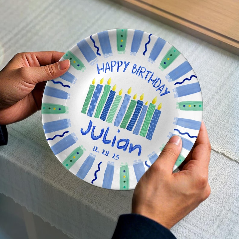 May include: A white plate with a blue and green design, featuring the text "HAPPY BIRTHDAY Julian" and the date "12. 28. 25". The plate has a border of blue and green stripes and wavy lines, and a row of candles.