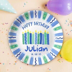 May include: A round, white birthday plate with blue and green accents. The plate features the text "HAPPY BIRTHDAY Julian" and a row of candles. The date "12. 28. 25" is printed below the name. Party supplies are scattered around the plate.