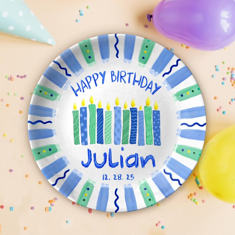 May include: A round, white birthday plate with blue and green accents. The plate features the text "HAPPY BIRTHDAY Julian" and a row of candles. The date "12. 28. 25" is printed below the name. Party supplies are scattered around the plate.