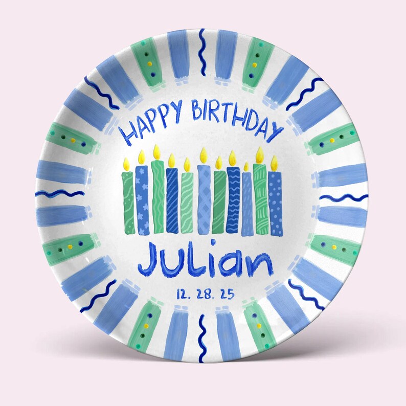 May include: A white ceramic plate with a blue and green painted design. The plate features the text "HAPPY BIRTHDAY" in blue, the name "Julian" in blue, and the date "12. 28. 25". The plate has a border of blue and green painted stripes and wavy lines.
