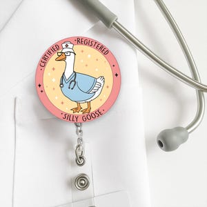 May include: A round badge reel featuring a cartoon goose wearing a nurse's uniform, glasses, and a hat. The badge reel has the words "CERTIFIED REGISTERED SILLY GOOSE" around the edge. A stethoscope is in the background.