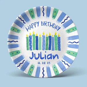 May include: A white ceramic plate with a blue and green painted design. The plate has the words "HAPPY BIRTHDAY" and the name "Julian" in blue. The plate also features candles and the date 12.28.25.