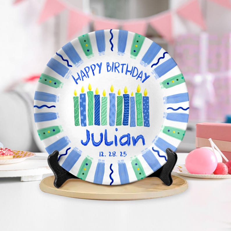 May include: A white plate with a blue and green striped border and the words "HAPPY BIRTHDAY" in blue. The plate features candles and the name "Julian" in blue, with the date 12.28.25. The plate is displayed on a black stand.