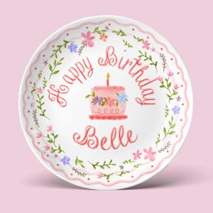 May include: A white ceramic plate with a pink floral border and the words "Happy Birthday Belle" in pink script. A pink cake with a lit candle is in the center, surrounded by flowers.
