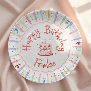 May include: A white, round birthday plate with a pink scalloped edge. The plate features the words "Happy Birthday Frankie" in red script, along with a pink cake with hearts and a lit candle. Colorful candles surround the edge.