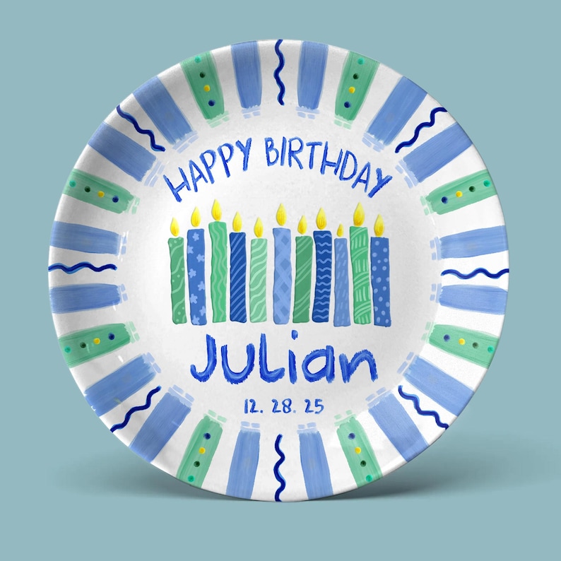 May include: A white ceramic plate with a blue and green painted design. The plate features the text "HAPPY BIRTHDAY" and "Julian" in blue, with a row of candles and the date 12.28.25.