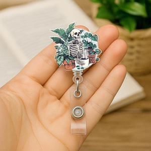 May include: A badge reel featuring a skeleton enjoying a cup of coffee amidst potted plants. The design includes a white skull, rib cage, and green foliage. The reel has a clear plastic strap and a metal clip.