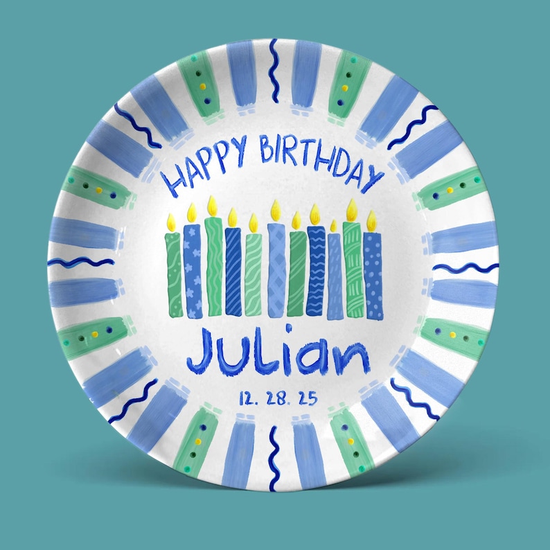 May include: A white ceramic plate with a blue and green border design. The plate features the text "HAPPY BIRTHDAY" and "Julian" in blue, with candles in the center. The date "12. 28. 25" is also included.