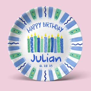 May include: A white ceramic plate with a blue and green painted border. The plate features the text "HAPPY BIRTHDAY" and "Julian" in blue, with the date "12. 28. 25" below. Ten candles are painted in the center.
