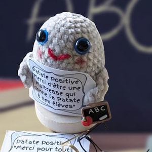 Puede incluir: Un personaje de patata de peluche hecho a mano con ojos azules, una sonrisa roja y un letrero que dice "Patate Positive". Una pequeña pizarra con "ABC" y una manzana roja está adjunta.