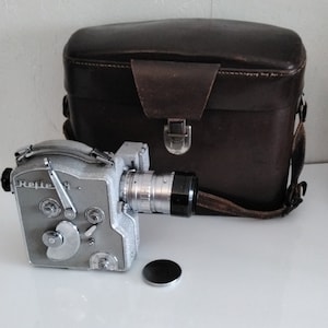 May include: A vintage silver and black Reflex 16mm movie camera with a lens and a black lens cap. A brown leather case with a handle and clasp is behind the camera. The camera is on a white surface.