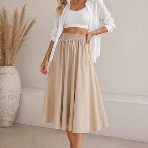 May include: A beige midi skirt with an elastic waistband is paired with a white crop top and a white button-down shirt. The outfit is completed with woven sandals. The image includes the text "Nosta Pattern" in the upper left corner.