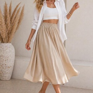 May include: A woman wearing a white crop top, a white button-down shirt, and a beige midi skirt. The skirt has an elastic waistband. The woman is also wearing sandals. The image has the text "Nosta Pattern" in the top left corner.