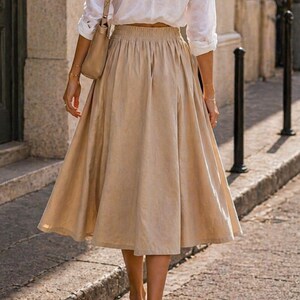 May include: A woman wearing a white button-down shirt and a beige skirt, walking on a cobblestone street. The skirt is a midi length with a gathered waist. She is carrying a beige purse and wearing sandals.