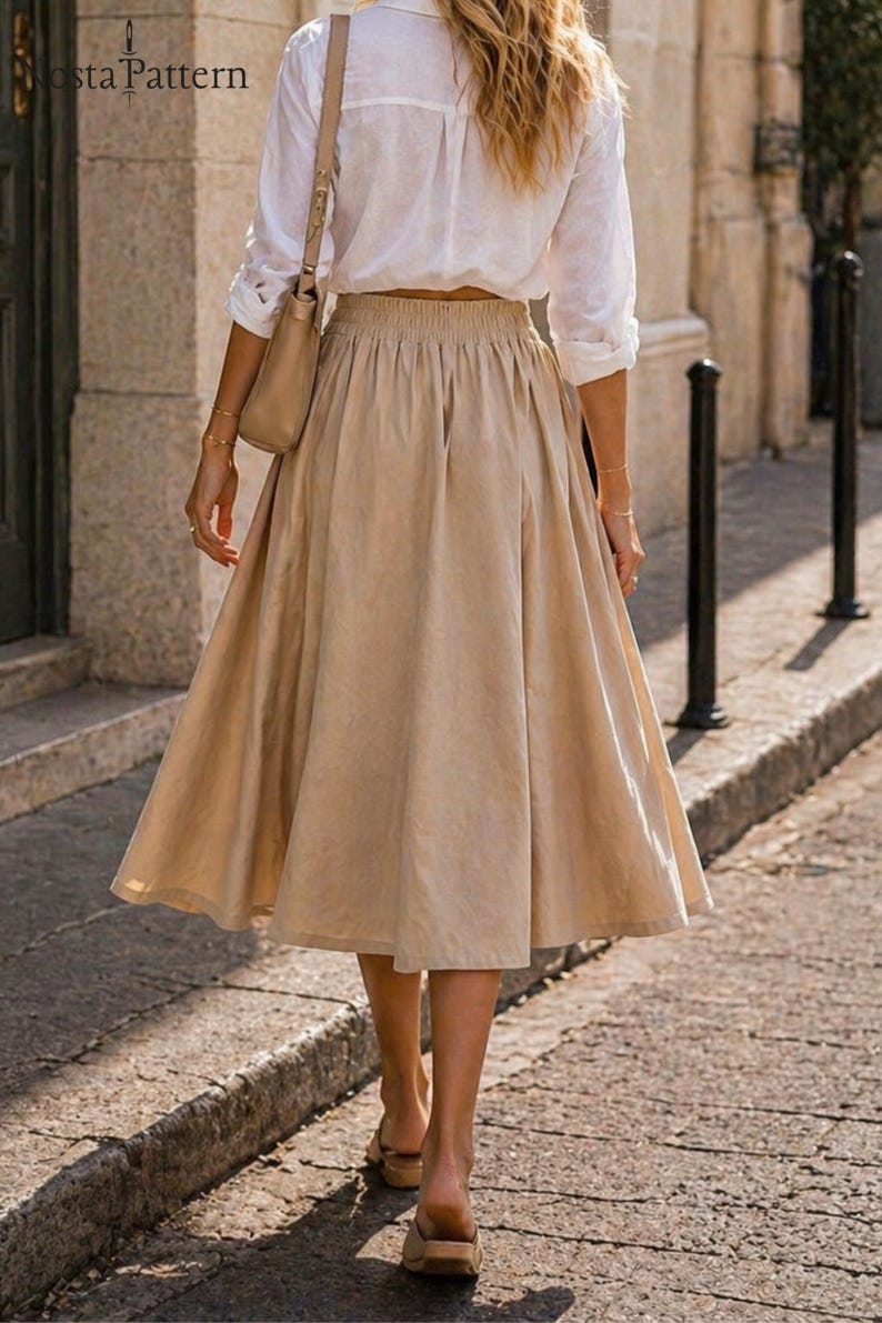 May include: A woman wearing a white button-down shirt and a beige skirt, walking on a cobblestone street. The skirt is a midi length with a gathered waist. She is carrying a beige purse and wearing sandals.