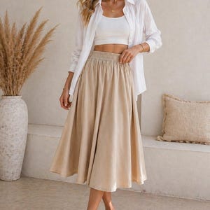 May include: A woman wearing a white crop top, white button-down shirt, and a beige midi skirt. The skirt has an elastic waistband. The image also shows a pair of woven sandals. The text "Nosta Pattern" is in the top left corner.