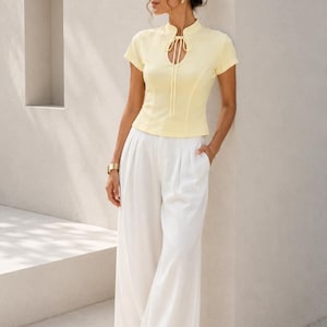 Peut inclure: Un haut jaune p&acirc;le &agrave; manches courtes avec un col officier, une encolure en trou de serrure avec un lien &agrave; nouer, et un pantalon blanc &agrave; jambes larges. Le haut est ajust&eacute; avec des coutures verticales et le pantalon a une taille pliss&eacute;e.