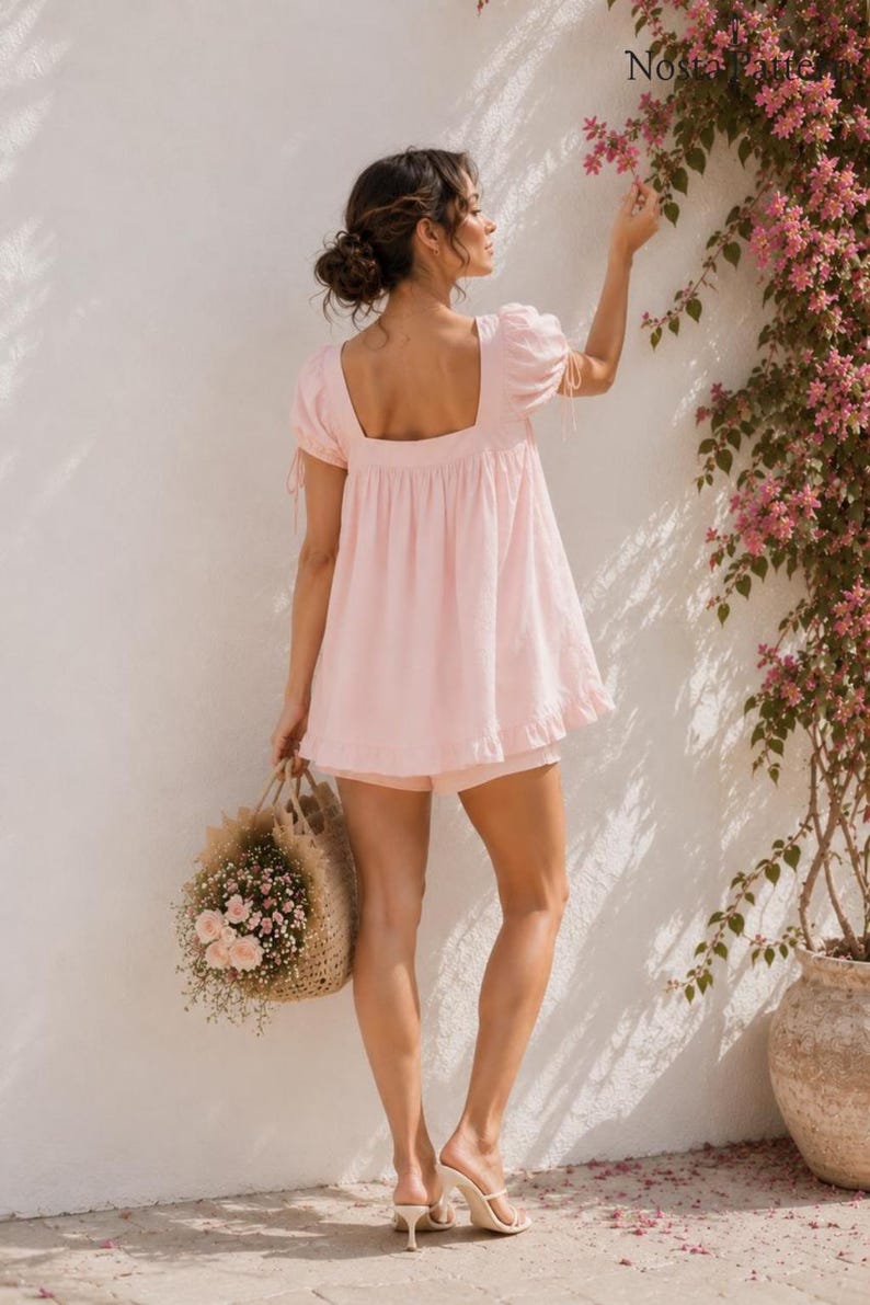 May include: A light pink short-sleeved top with a square neckline, ruffled hem, and tie details on the sleeves. The garment is paired with matching light pink shorts. The text Nosta Pattern is visible in the upper right corner of the image.