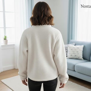 Puede incluir: Un jersey de forro polar cremoso y difuso con cuello alto. La vista trasera muestra la textura suave de la tela. La persona lleva pantalones negros. El fondo incluye una ventana, un sof&aacute; y el logotipo "Nosta Pattern".