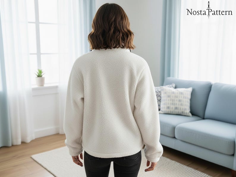Puede incluir: Un jersey de forro polar cremoso y difuso con cuello alto. La vista trasera muestra la textura suave de la tela. La persona lleva pantalones negros. El fondo incluye una ventana, un sof&aacute; y el logotipo "Nosta Pattern".