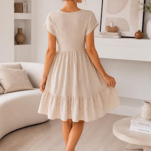 May include: A beige knee-length dress with short sleeves, a fitted bodice, and a flared skirt featuring a horizontal striped pattern at the hem. The garment has a V-neckline and a textured fabric finish, shown from the back view.