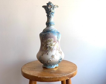 May include: A tall, blue and white porcelain vase with a floral design and gold accents. The vase is sitting on a wooden stool.