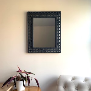 May include: A black rope mirror with a decorative woven frame hangs on a white wall above a wooden stool and a white tufted armchair.