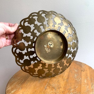 Może przedstawiać: Dekoracyjna miska z mosiądzu z wyciętym wzorem. Miska ma okrągły kształt i falisty brzeg. Miska jest wykonana z mosiądzu i ma złote wykończenie. Miska stoi na drewnianej powierzchni.