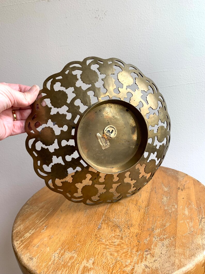 Może przedstawiać: Dekoracyjna miska z mosiądzu z wyciętym wzorem. Miska ma okrągły kształt i falisty brzeg. Miska jest wykonana z mosiądzu i ma złote wykończenie. Miska stoi na drewnianej powierzchni.