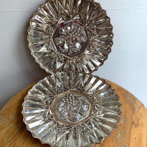 May include: Two silver-colored glass plates with a raised design of fruit, including pears and grapes. The plates have a scalloped edge and a clear glass base.