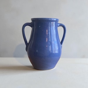 May include: A blue ceramic vase with two curved handles. The vase has a rounded body and a slightly flared rim. The color is a solid, medium blue with a glossy finish. The vase is likely handmade and suitable for displaying flowers.