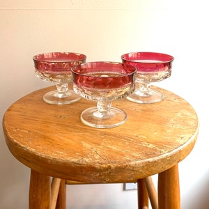 May include: Three clear glass dessert dishes with a ruby red rim. The dishes have a scalloped edge and a pedestal base.
