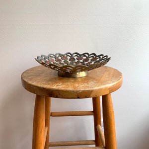 Może przedstawiać: Dekoracyjna misa z mosiądzu z falistym brzegiem i wzorem kwiatowym. Misa stoi na drewnianym stołku.