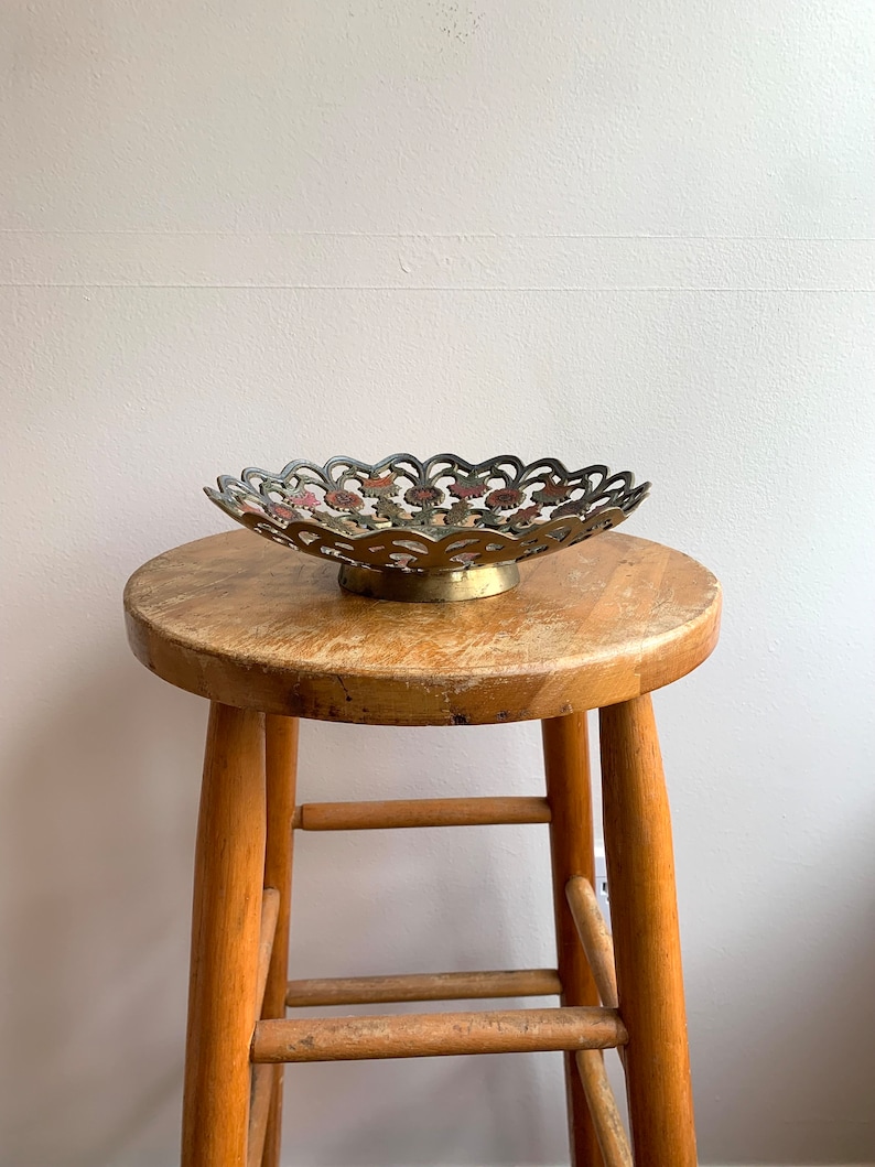 Może przedstawiać: Dekoracyjna misa z mosiądzu z falistym brzegiem i wzorem kwiatowym. Misa stoi na drewnianym stołku.