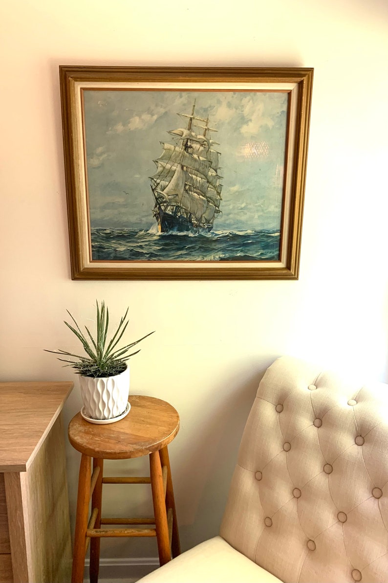 May include: A framed vintage print of a sailing ship with white sails against a blue sky and ocean. The ship is in the center of the image and is surrounded by water. The frame is gold and has a white mat.