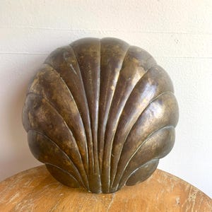 May include: A large, antique-style, bronze-colored seashell wall hanging. The shell has a detailed, fan-like design with vertical ridges. The surface has a weathered appearance, suggesting age and a vintage aesthetic. The shell is resting on a wooden surface.