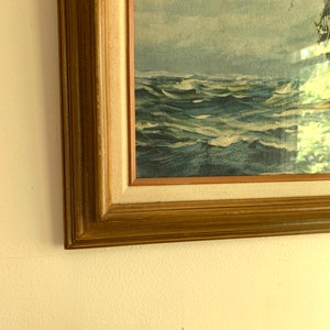 May include: A framed painting of a ship sailing on a stormy sea. The painting is in a wooden frame with a gold finish. The sea is a deep blue with whitecaps and the sky is a light blue with white clouds.