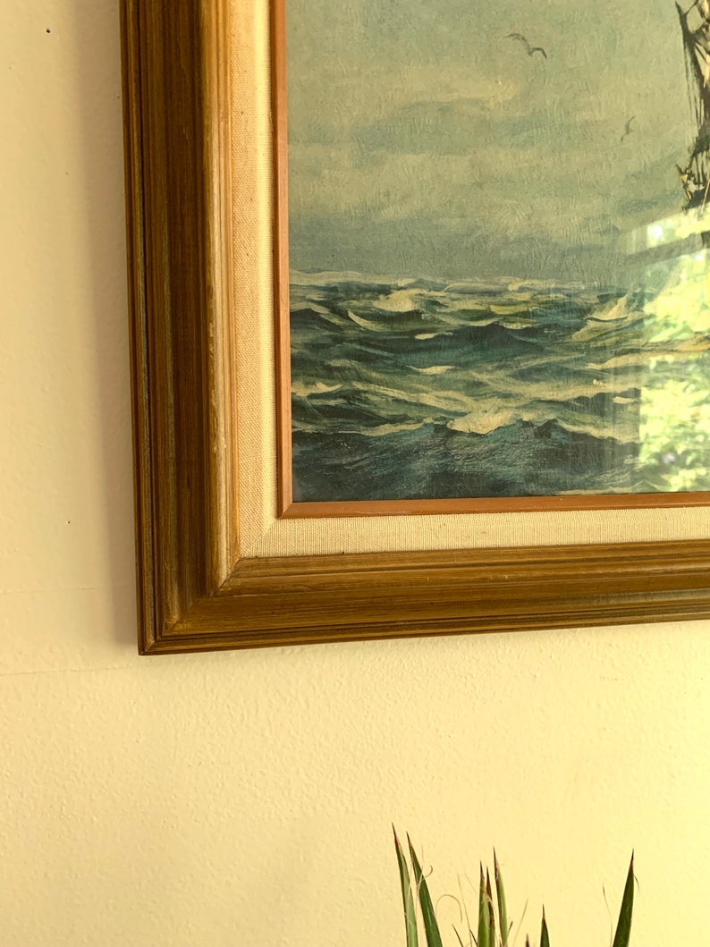 May include: A framed painting of a ship sailing on a stormy sea. The painting is in a wooden frame with a gold finish. The sea is a deep blue with whitecaps and the sky is a light blue with white clouds.