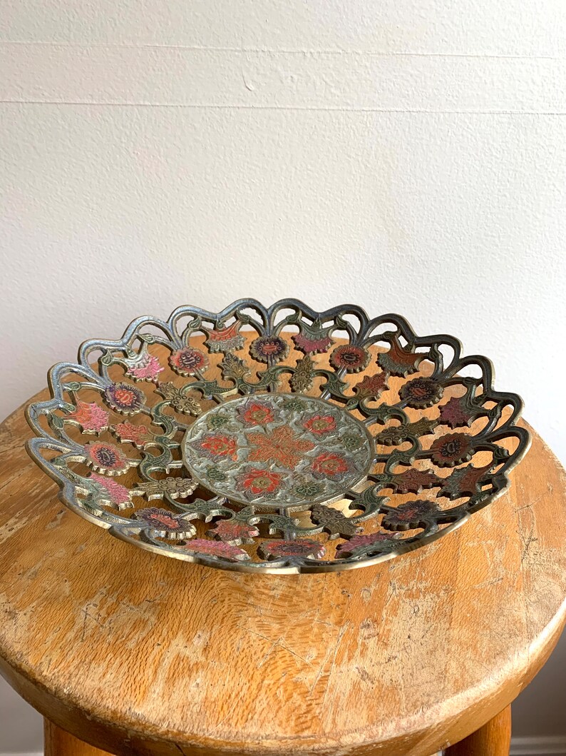 Może przedstawiać: Miska mosiężna z misternym wzorem kwiatowym w odcieniach r&oacute;żowego, zielonego i złotego. Miska ma falisty brzeg i wypukłe centrum.