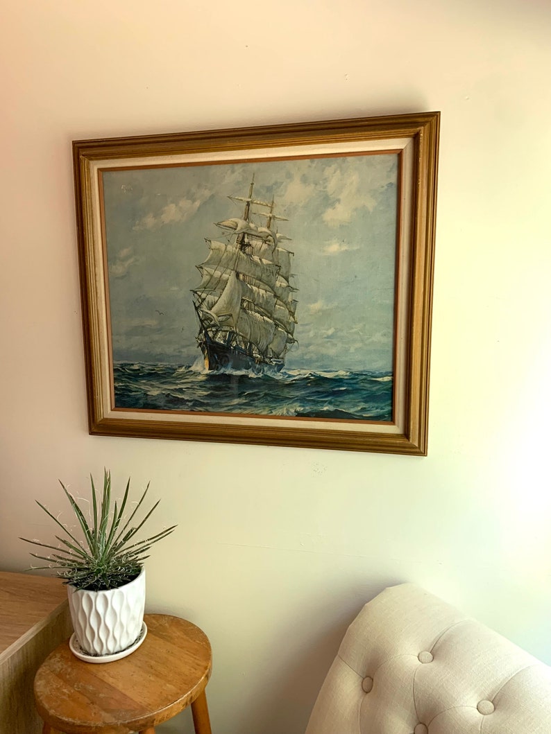 May include: A framed painting of a tall ship sailing on a blue ocean with white clouds in the sky. The ship has white sails and a brown hull. The frame is gold.