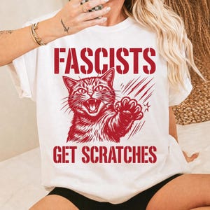 Peut inclure: T-shirt blanc avec un motif rouge d'un chat griffant et le texte "Fascists Get Scratches". L'illustration du chat est détaillée avec une gueule ouverte et une patte levée. Le texte est en gras, sans empattement.