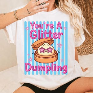Puede incluir: Camiseta blanca con un gráfico de una empanadilla con gafas de sol en forma de estrella dentro de una vaporera. El texto "You're A Glitter Dumpling" es en rosa, con rayas verticales azules y estrellas rosas.