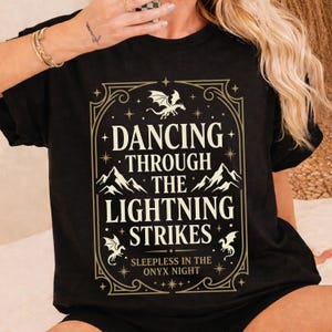 Könnte beinhalten: Schwarzes T-Shirt mit einem goldfarbenen, gerahmten Design. Das Design enthält den Text "DANCING THROUGH THE LIGHTNING STRIKES" mit Drachen- und Bergillustrationen. Der Satz "SLEEPLESS IN THE ONYX NIGHT" ist ebenfalls enthalten.