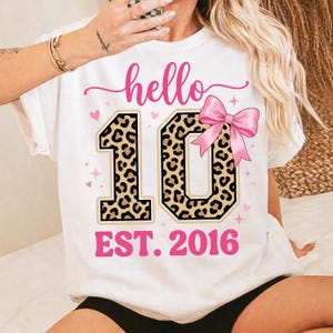 Könnte beinhalten: Weißes T-Shirt mit einem rosa "hello"-Schriftzug und einer großen "10" in Leopardenmuster. Eine rosa Schleife befindet sich auf der rechten Seite der "10". Der Text "EST. 2016" steht unter der Zahl. Rosa Herzen und Sterne sind um das Design verstreut.