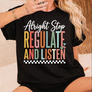 Puede incluir: Una camiseta negra de manga corta con el texto blanco Alright Stop y el texto apilado de colores REGULATE AND LISTEN en amarillo, rojo, naranja, azul y rosa. El diseño incluye corazones pequeños y un borde a cuadros blanco y negro.