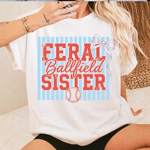 Op de afbeelding: Wit T-shirt met de tekst "FERAL Ballfield SISTER" in rood, omlijnd met wit, op een lichtblauwe gestreepte achtergrond. Een honkbalafbeelding staat onderaan. Een rode strik staat rechtsboven.