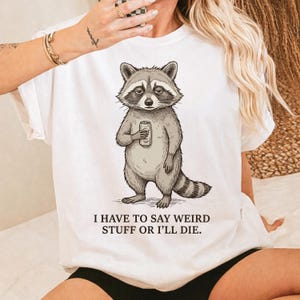 May include: White t-shirt featuring a cartoon raccoon holding a can. The raccoon has a sad expression. The text "I HAVE TO SAY WEIRD STUFF OR I'LL DIE" is printed below the raccoon illustration.