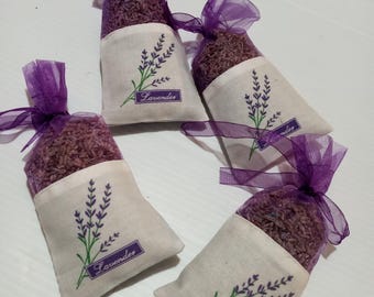 May include: Four small, white fabric sachets filled with dried lavender. Each sachet has a purple ribbon tie and a lavender sprig design with the word "Lavender" printed on it. The sachets are arranged on a white surface.