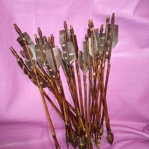 May include: A bundle of handcrafted arrows with wooden shafts, feathered fletching, and stone arrowheads. The arrows are arranged against a pink background, showcasing their natural materials and traditional design.