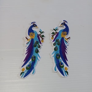 May include: Two embroidered peacock patches. Each patch features a peacock with blue, purple, and yellow details. The peacocks are perched on branches with green leaves and yellow flowers. The patches have a white border.